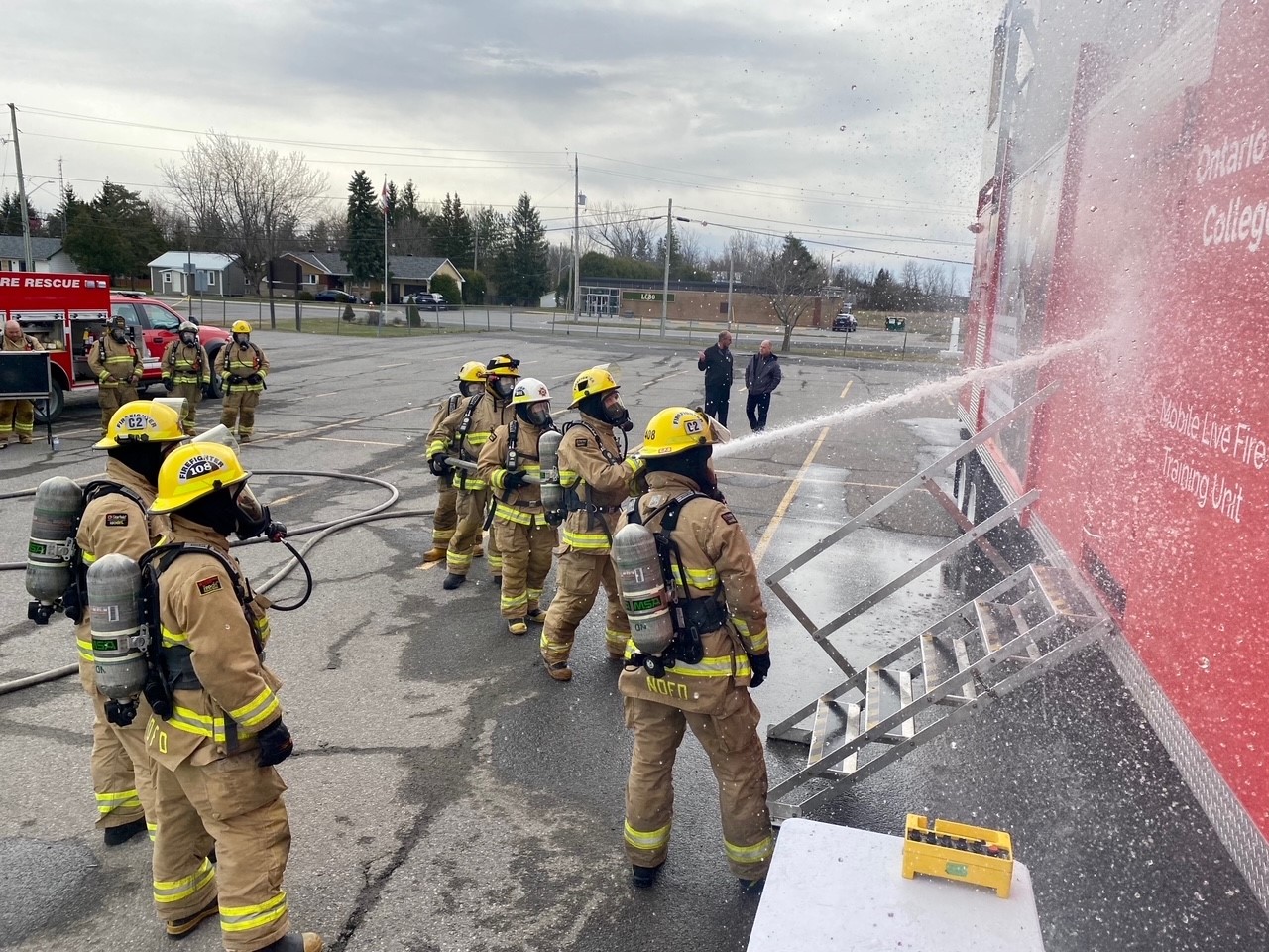 firefighters live hose spraying training onto side of red fire training trailer